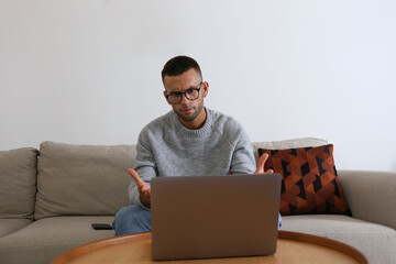 Frustrated man wearing glasses looking at the screen of his laptop with anger. Freelancer working on a couch in his living room. Close up, copy space, white wall background.
