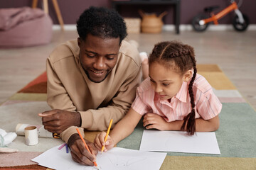 Portrait of Black father and daughter drawing pictures together laying on floor at home and smiling