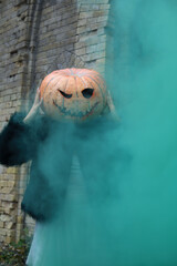 Halloween holiday. a girl in a green fur coat and with an orange pumpkin instead of a head. green smoke around. in the background there is a brick old building