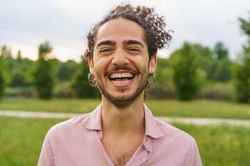 Outdoor portrait of a young man with curly hair tied back, laughing heartily with eyes squinted. He...