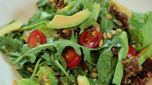 Salad rotates on a white plate: lettuce, avocado, cherry tomatoes, arugula, pumpkin seeds.