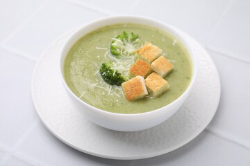 Delicious broccoli cream soup with croutons and cheese on white tiled table, closeup