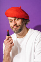 Funny middle aged man wearing red beret is holding lipstick in his hand against purple background