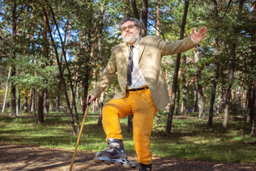 Funny old man is roller skating in the park. An active grandfather with a walking stick dances and...