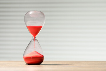 Hourglass with red flowing sand on wooden table against light background, space for text