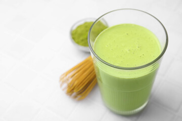Glass of tasty matcha smoothie, powder and bamboo whisk on white tiled table, closeup. Space for text