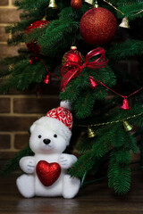 Snow-white teddy bear with heart in hands on the background of a Christmas tree. New Year's Card