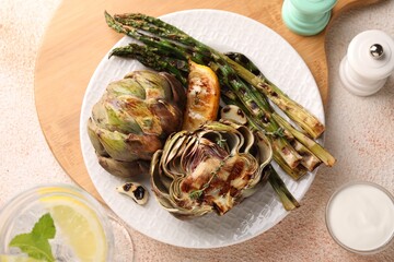 Tasty grilled artichokes and asparagus served on beige textured table, flat lay