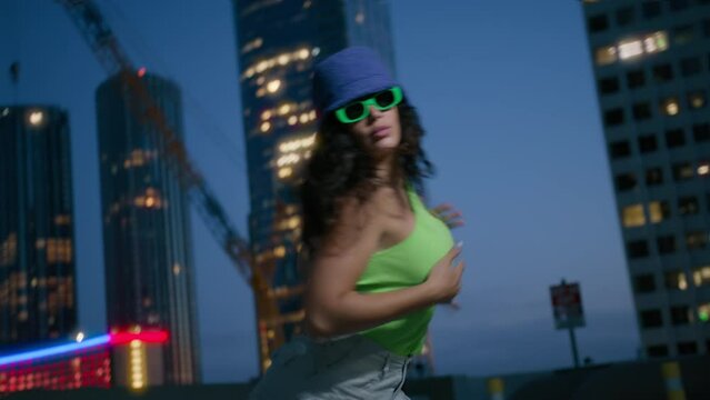 Cool girl dancer dancing in night city. Black young girl with long curly hair moves rhythmically. Sexy fashionable dancer in trendy bucket cup and bright green glasses moves sensually urban downtown