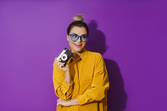 Cheerful girl wearing eyeglasses holding vintage film camera on purple background