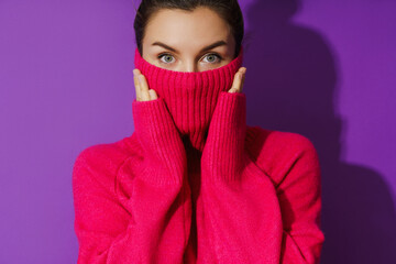 Young woman is hiding her face inside a warm and cozy polo neck sweater against purple background