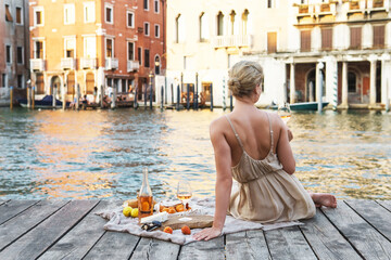 Lovely young woman picturesque picnic on the wooden gondola dock with rose wine, fruits and snack on wooden pier