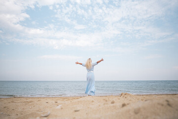 sea, sky, clouds, sunset, ocean, blonde woman in a floating dress stands with her back to the...