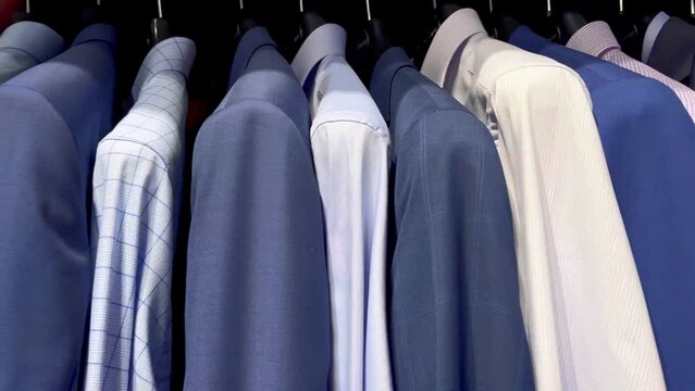 Fashionable men's shirts and jackets hang on hangers in a modern shopping mall. Shopping mall boutique filled with fashion tailoring, multiple racks with fashionable formal wear. Buying clothes.