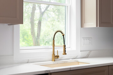 A gold kitchen faucet detail with a gold sink, brown cabinets, and a white marble countertop.