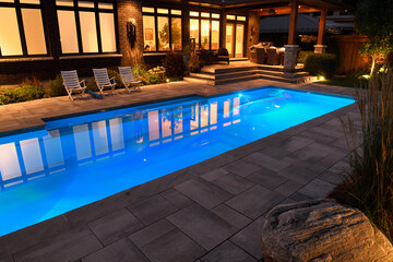 Home with outdoor patio and lights on at night and blue lit swimming pool with stone deck - obrazy, fototapety, plakaty