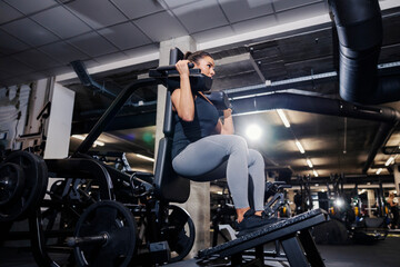 A strong woman is doing exercises for legs and shoulders on a workout machine in a gym.