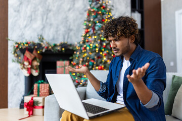 Upset man looking at laptop screen sitting at home in living room on Christmas, hispanic man frustrated writing online notification email to mail, near Christmas tree on New Year holidays.