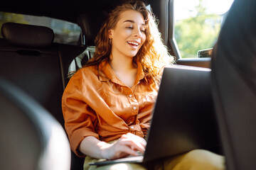 A young curly woman with a laptop sitting in the back seat of a car. Beautiful woman uses laptop in car. Remote work, freelance.