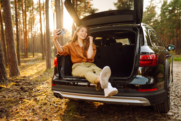 Stylish woman takes a selfie, communicates via video call from a smartphone in the trunk of a car at sunset. Autumn trip. A young woman uses the phone and travels by car.
