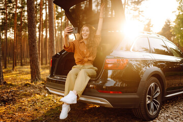 Stylish woman takes a selfie, communicates via video call from a smartphone in the trunk of a car at sunset. Autumn trip. A young woman uses the phone and travels by car.