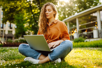 Joyful woman with laptop outdoors. A young woman sits on a green lawn and works with a laptop. Concept of blogging, freelancing, relax.