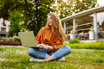 Joyful woman with laptop outdoors. A young woman sits on a green lawn and works with a laptop. Concept of blogging, freelancing, relax.