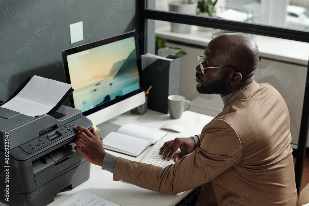 Poster young confident man in beige blazer pressing start button on panel of xerox machine while making som