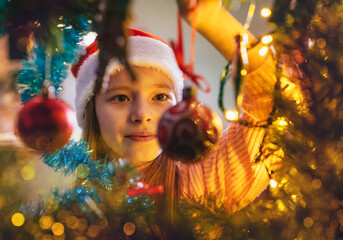 Cute little girl in red Santa hat hanging a magic tree decoration ball on the Christmas fir-tree on...