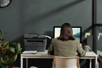 Rear view of young brunette secretary sitting on chair in front of computer screen in office and searching for online information in the internet
