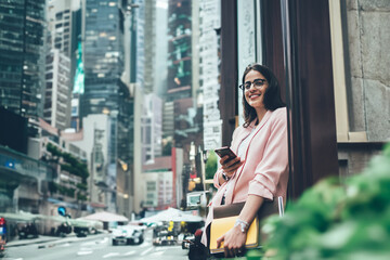 Cheerful busy woman with smartphone and papers standing next to building