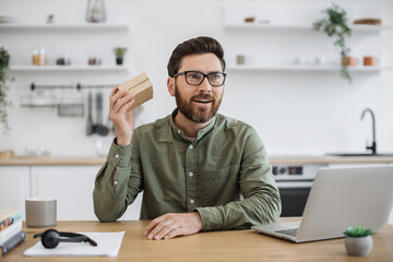 Caucasian bearded man in eyeglasses and looking at small cardboard parcel with focused facial expression. Social media influencer streaming unpacking process of online order from home.