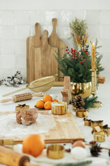 Gingerbread dough with flour on wooden board, rolling pin, golden metal cutters, cooking spices and festive decorations on countertop in modern white kitchen. Making christmas gingerbread cookies