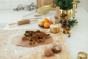 Fototapeta na wymiar Gingerbread dough with festive golden metal cutters on wooden board with flour, cooking spices, festive decorations in modern white kitchen. Making christmas gingerbread cookies