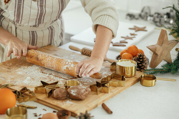 Woman making christmas gingerbread cookies in modern white kitchen close up. Hands kneading gingerbread dough with rolling pin, golden metal cutters, cooking spices, festive decorations