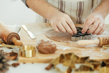 Man making christmas gingerbread cookies close up in modern white kitchen. Hands cutting gingerbread dough with festive golden metal cutters with cooking spices and decorations
