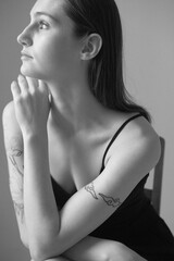 Black and white portrait of a brunette girl without make up on a grey backgroung in a black summer clothes. Woman is sitting on a chair
