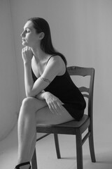 Black and white portrait of a brunette girl without make up on a grey backgroung in a black summer clothes. Woman is sitting on a chair