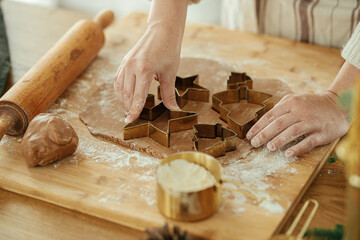 Woman making christmas gingerbread cookies, atmospheric time. Hands cutting gingerbread dough with festive golden cutters on rustic table with holiday decorations, close up