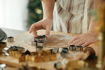 Hands cutting gingerbread dough with festive cutters on rustic table with holiday decorations against stylish christmas tree. Man making christmas gingerbread cookies, atmospheric holiday time
