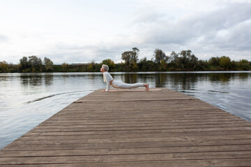 Obraz premium A mature woman with a short haircut in a white suit is doing yoga on the riverbank. Bhujangasana. Upward Dog