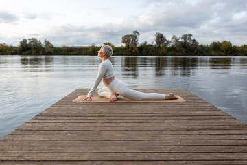 A mature woman with a short haircut in a white suit is doing yoga on the riverbank. Eka Pada Rajakapotasana