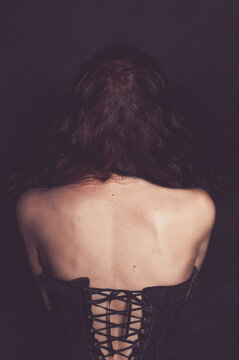 The bare back of a girl dressed in a black corset, a pearl necklace around her neck, and dark curly hair. Gothic clothing style.