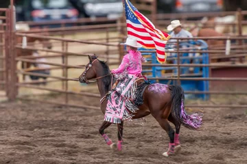 Fotobehang Paardensport A cowgirl is riding a horse at a rodeo in a dirt arena. She is holding an American flag. The horse is brown and has sequence on its back . She has a pink blouse and a white hat.   © Timothy