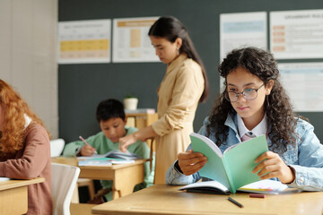 Clever schoolgirl in eyeglasses looking through her notes in copybook against teacher checking home assignment of one of classmates