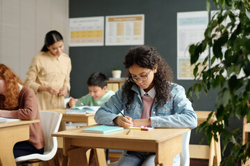 Diligent multiethnic schoolgirl making notes in copybook during individual work at lesson while teacher checking assignment of schoolboy