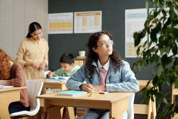 Fototapeta premium Cute pensive secondary school learner looking through window of classroom while sitting by desk and writing down essay at lesson