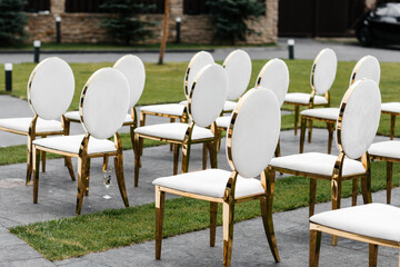 Luxury white-golden chairs on wedding ceremony outdoors. Festive decorations. Empty rows armchairs for guests. Outgoing wedding ceremony in the open air. Decor Studio.
