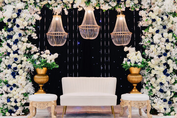 A beautiful decorated place for the wedding ceremony of the bride and groom in oriental style. Wedding arch made of white and blue fresh flowers. Beautiful decorative chandeliers and a white bench - obrazy, fototapety, plakaty