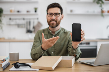 First person view of male influencer sharing feedback with followers while live streaming unpacking of modern smartphone. Bearded man sitting at desk and pointing with hand on new cell phone.